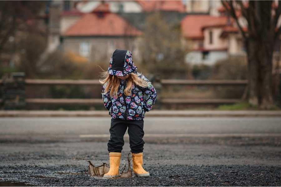 Little girl with long blond hair, a hood on her head, yellow boots, steps into a puddle until the water splashes